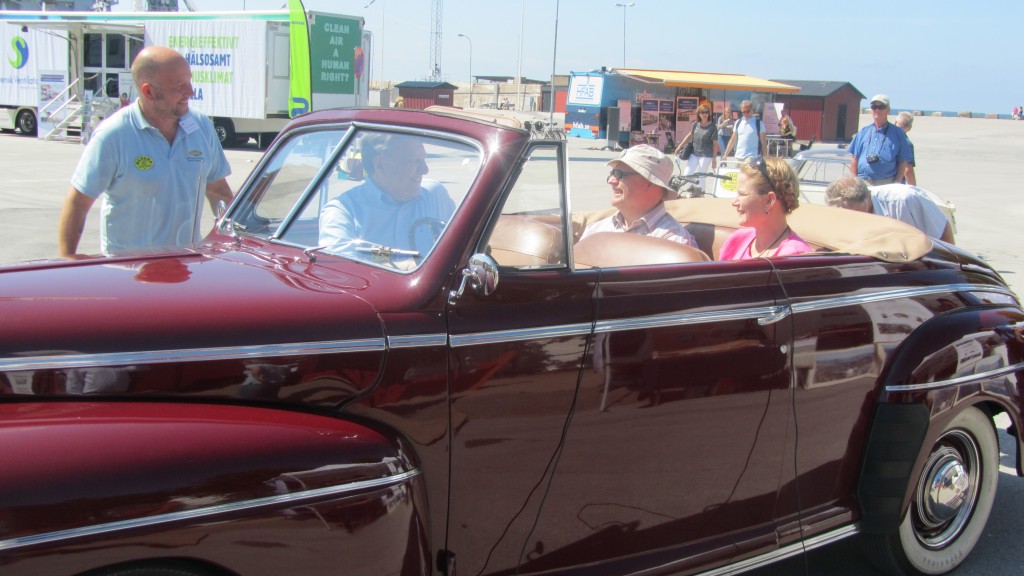 I baksätet på en Ford cabriolet från 50-talet sitter en man och en kvinna. I framsätet sitter en man vänd mot dem.